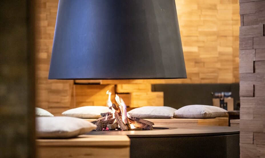 Feuerstelle mit brennendem Holz unter einer schwarzen Metallhaube, umgeben von Kissen