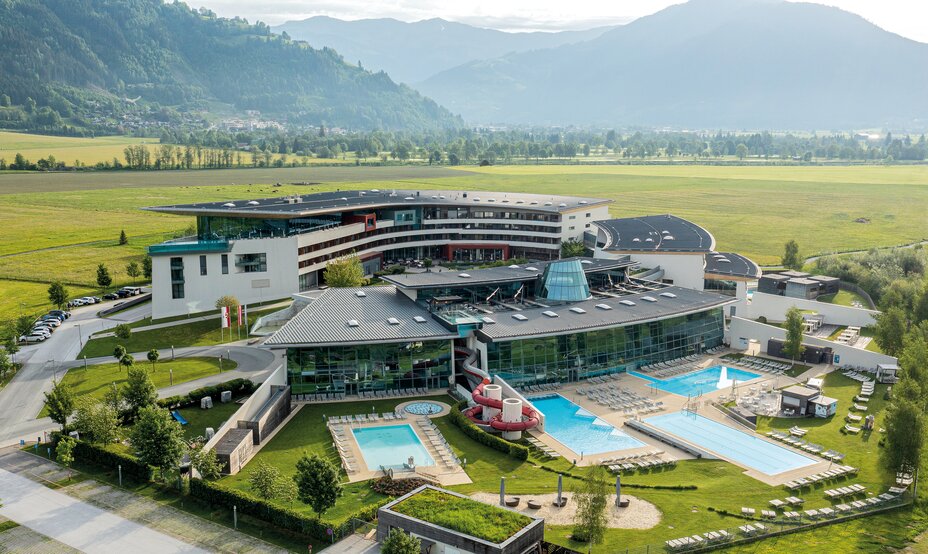 Hotelanlage mit Pools, Rutsche und grüner Wiese vor Bergkulisse