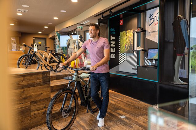 Mann schiebt E-Mountainbike in Fahrradverleih mit Aufschrift Rent