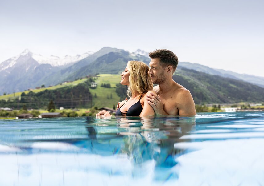 Paar im Infinity-Pool vor Bergpanorama, Frau mit blonden Haaren, Mann ohne Oberteil