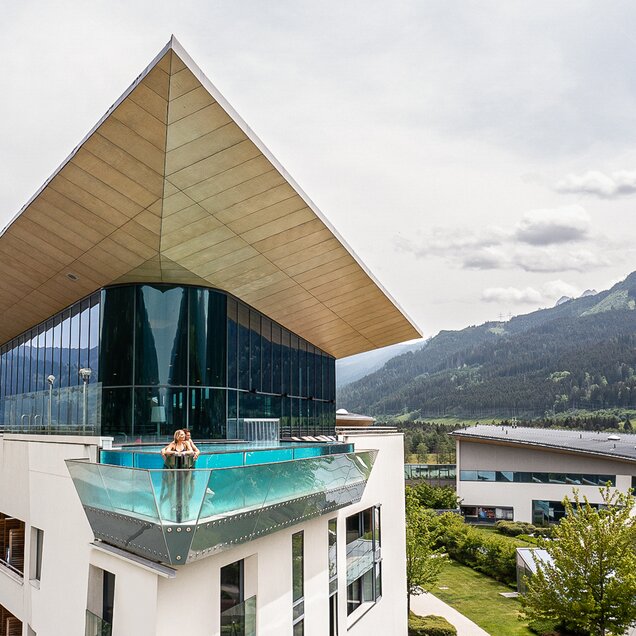 Woman in skyline pool on hotel roof with glass front in front of mountain panorama