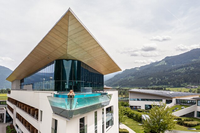 Frau im Skylinepool auf Hoteldach mit Glasfront vor Bergpanorama
