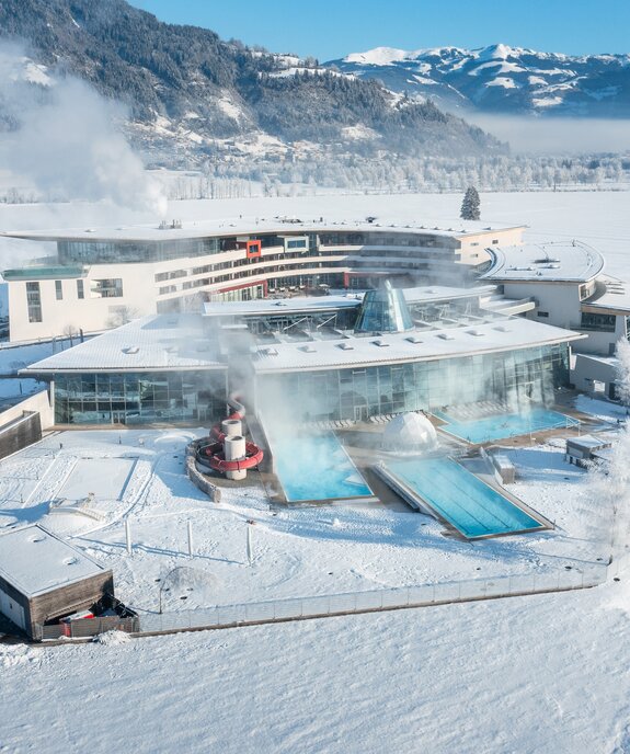 Luftaufnahme einer Therme im Winter mit schneebedeckter Landschaft und Bergen im Hintergrund