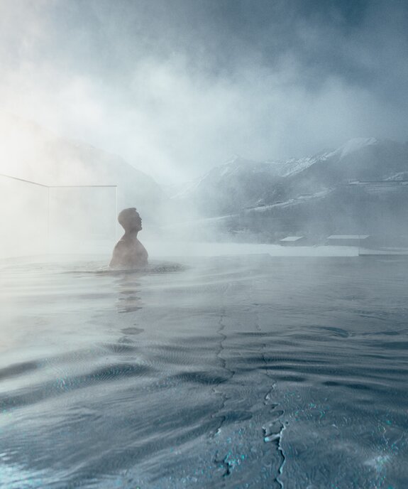Person im dampfenden Glas-Panoramapool vor verschneiter Winterlandschaft
