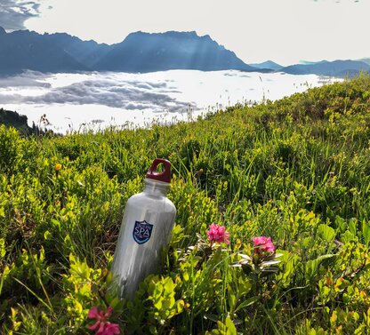 Trinkflasche in Almwiese mit rosa Blumen vor Bergpanorama mit Wolkenmeer