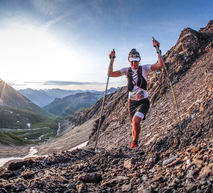 Läuferin mit Stöcken auf steinigem Bergpfad vor Alpenpanorama