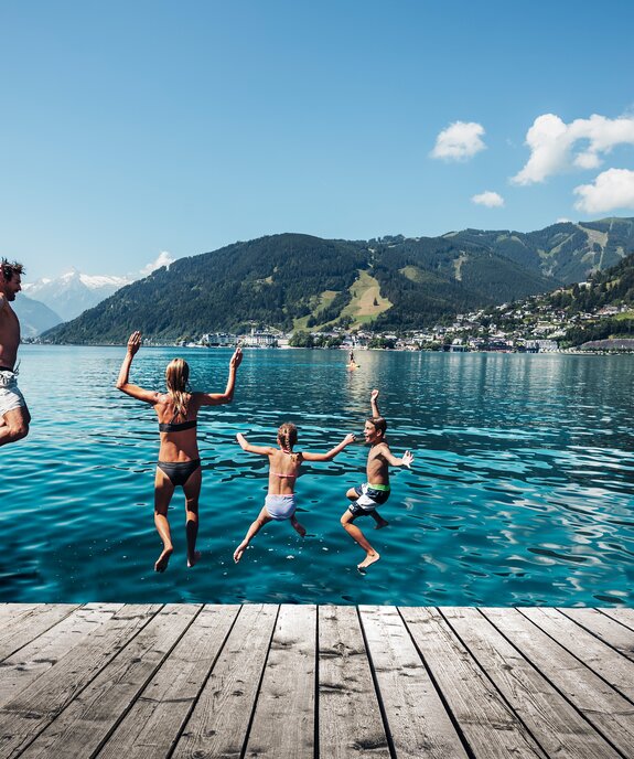 Familie springt von Holzsteg in türkisfarbenen See vor Bergkulisse unter blauem Himmel