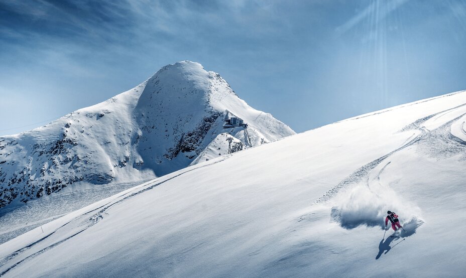 Skifahrer fährt Tiefschneehang hinab, im Hintergrund schneebedeckter Berg unter blauem Himmel