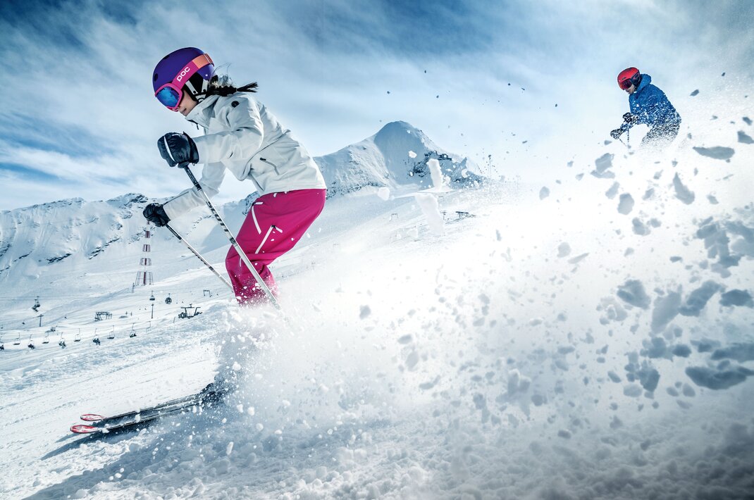Skifahrerin in weißer Jacke und pinker Hose fährt Piste hinunter, Kitzsteinhorn im Hintergrund