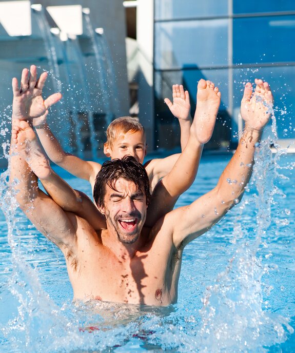 Man with child on shoulders in swimming pool, both laughing and raising arms