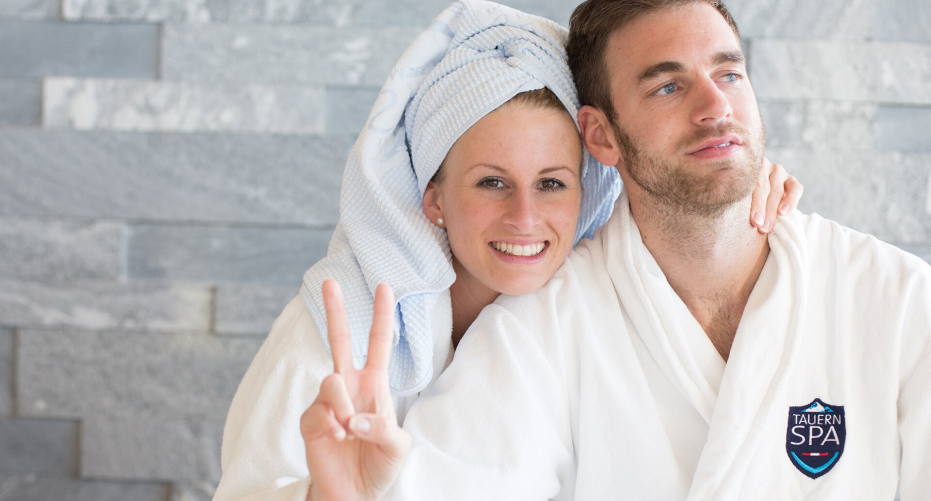Smiling couple in bathrobes, woman with towel on head showing victory sign