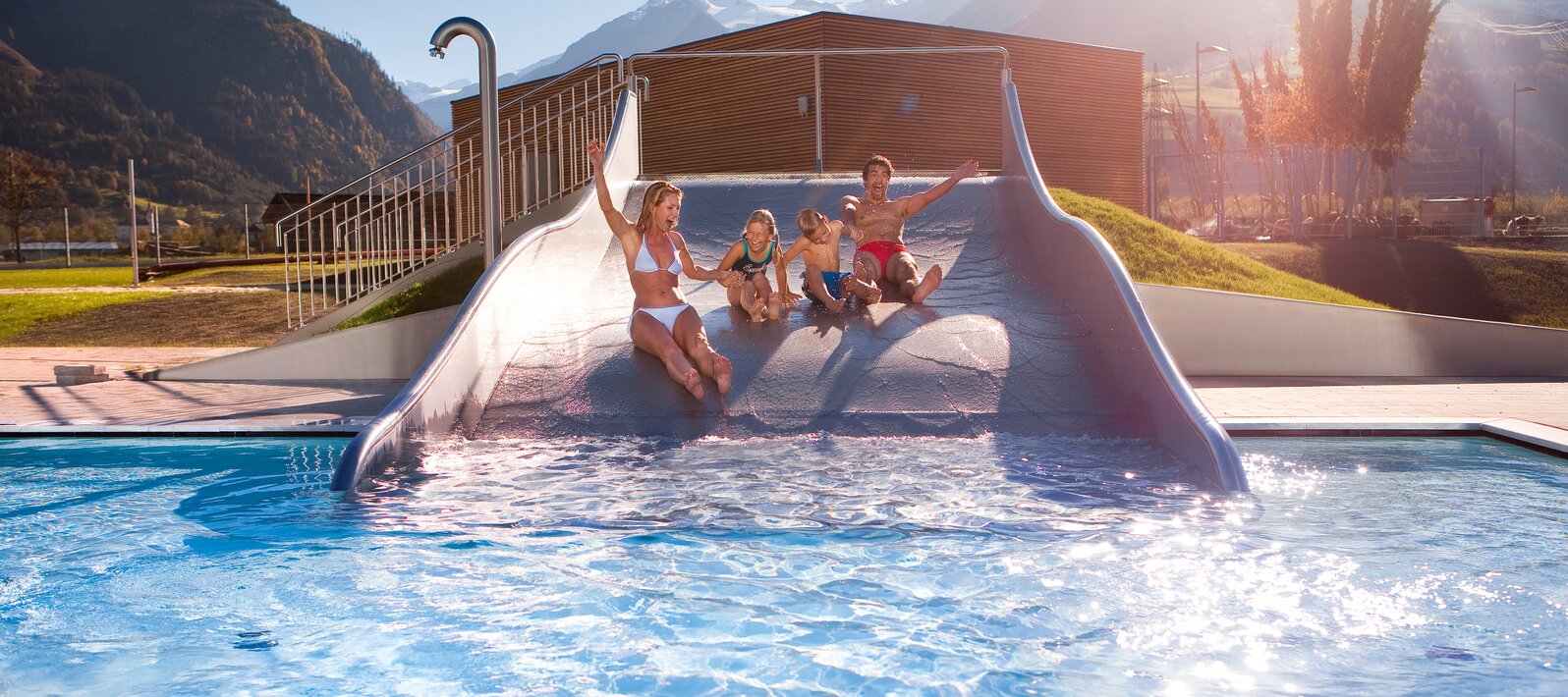 Familie rutscht auf einer Wasserrutsche in ein Schwimmbad vor Bergkulisse