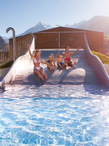 Familie rutscht auf einer Wasserrutsche in ein Schwimmbad vor Bergkulisse