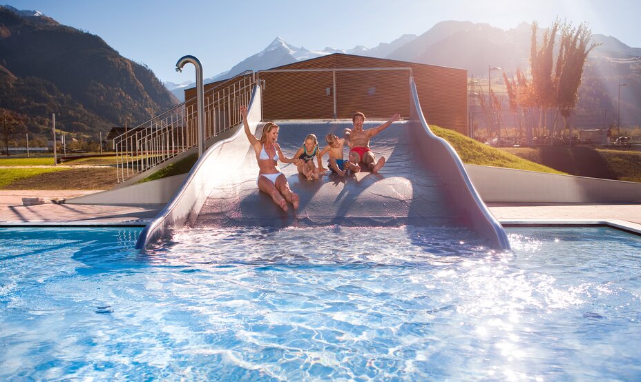 Familie rutscht auf einer Wasserrutsche in ein Schwimmbad vor Bergkulisse