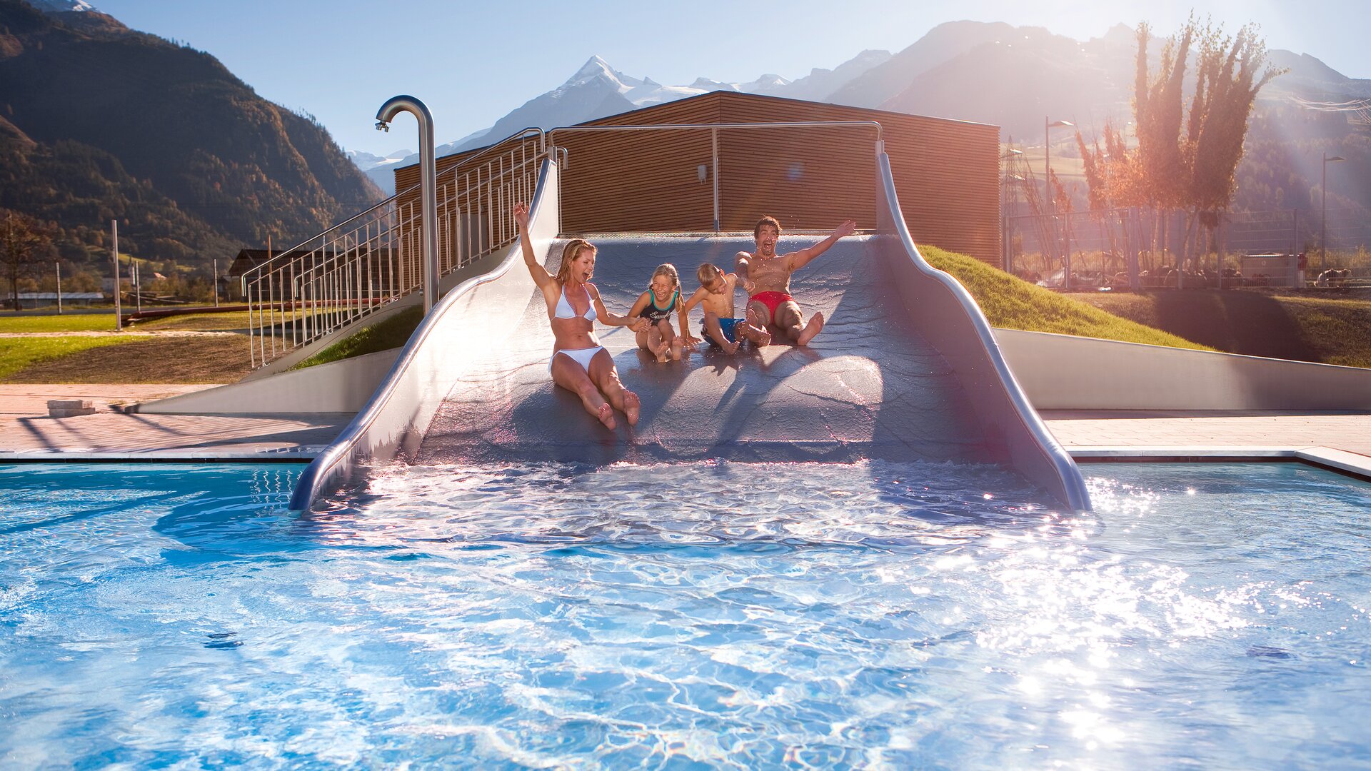 Familie rutscht auf einer Wasserrutsche in ein Schwimmbad vor Bergkulisse