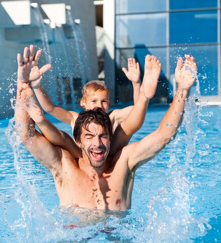 Man with child on shoulders in swimming pool, both laughing and raising arms