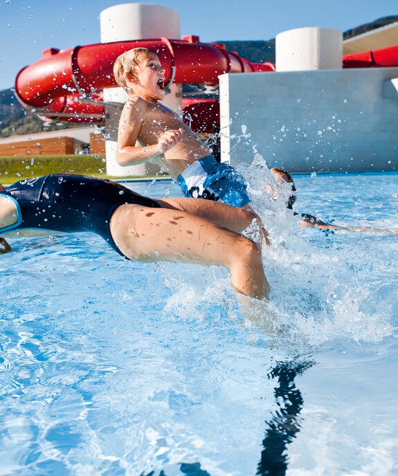 Kinder planschen im Freien in einem Schwimmbecken vor einer roten Wasserrutsche