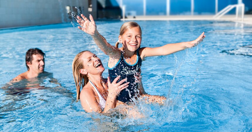 Mutter hält lachendes Mädchen im Schwimmbecken, spritzendes Wasser