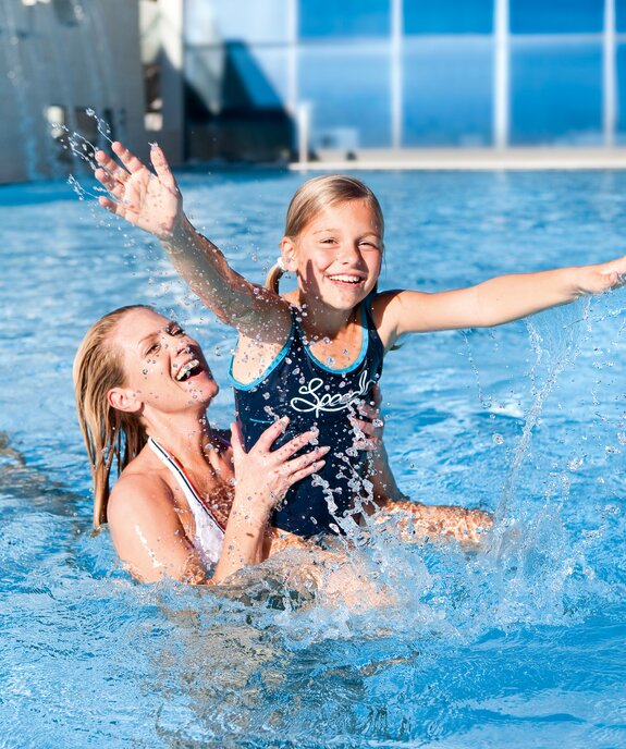 Mutter hält lachendes Mädchen im Schwimmbecken, spritzendes Wasser