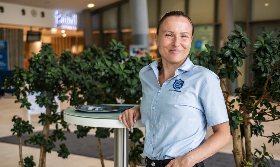 Frau in hellblauem Hemd mit Logo lehnt an rundem Tisch vor grünen Pflanzen