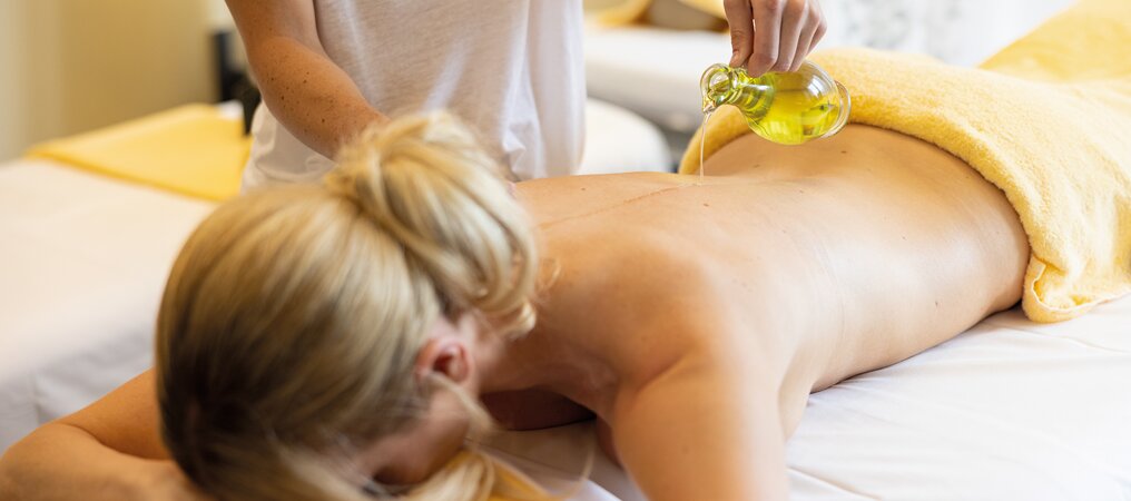 Frau liegt auf Bauch, während Masseurin Öl auf ihren Rücken gießt